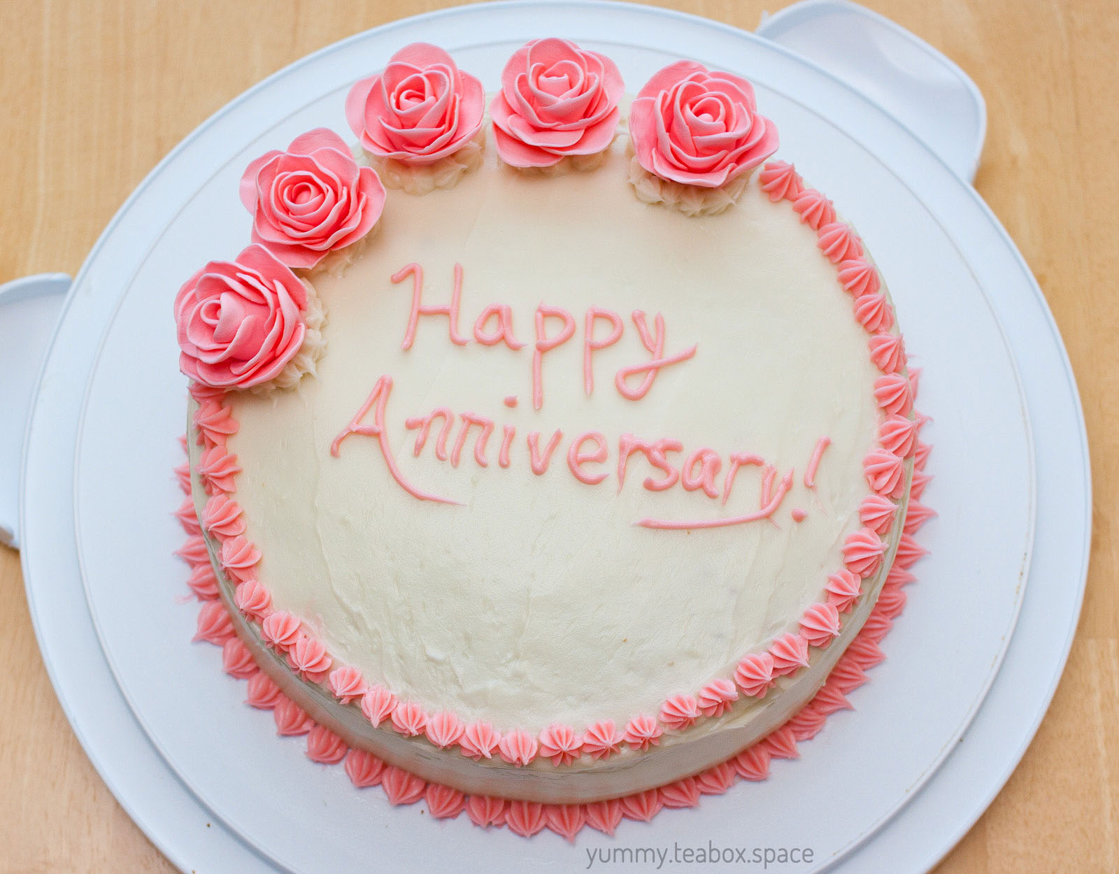 Round white cake with a pink border and five pink roses on the top left. The cake says Happy Anniversary.