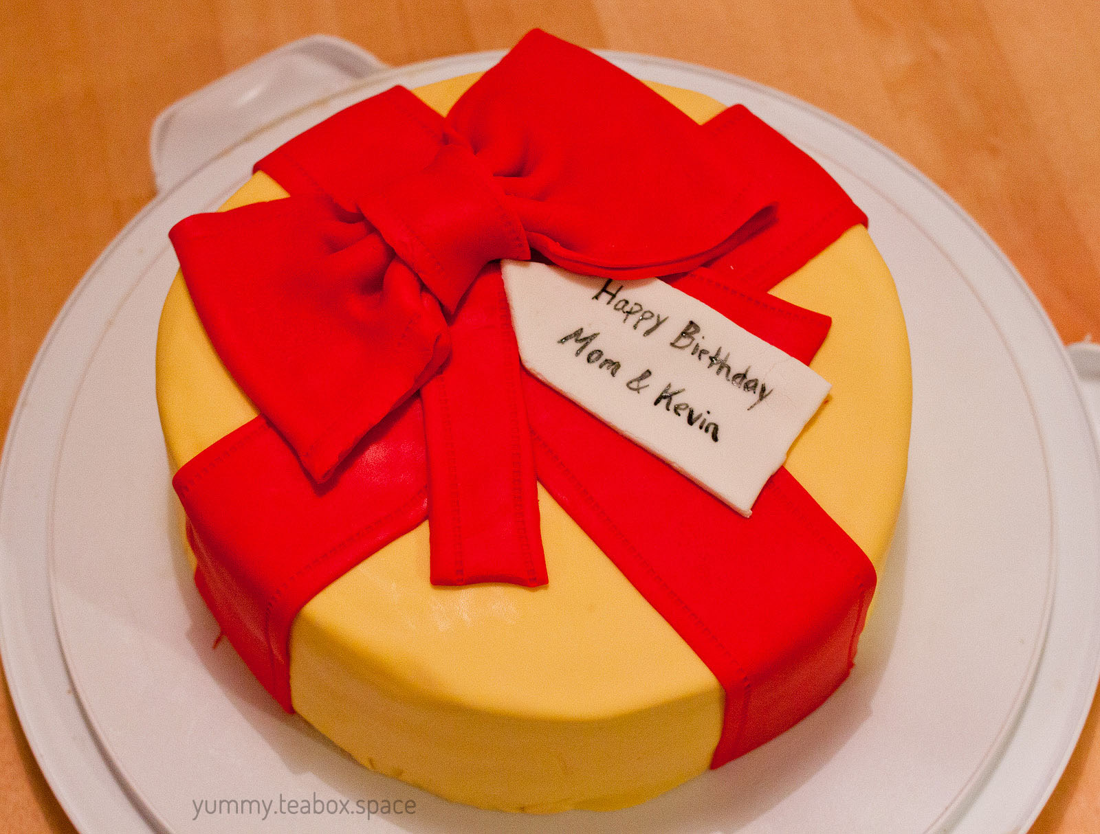 A round cake that looks like a yellow present with red ribbon, a red bow, and a gift tag that says "Happy Birthday Mom & Kevin"