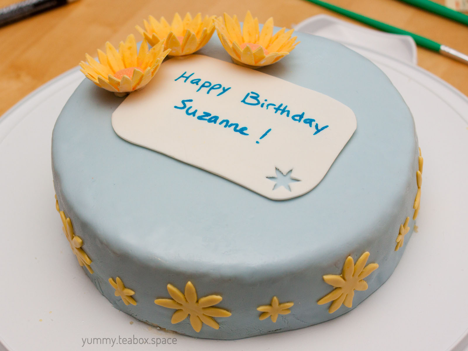 Round cake covered in blue fondant with yellow flowers on the top left and yellow flowers around the edge. It says Happy Birthday Suzanne in the center.