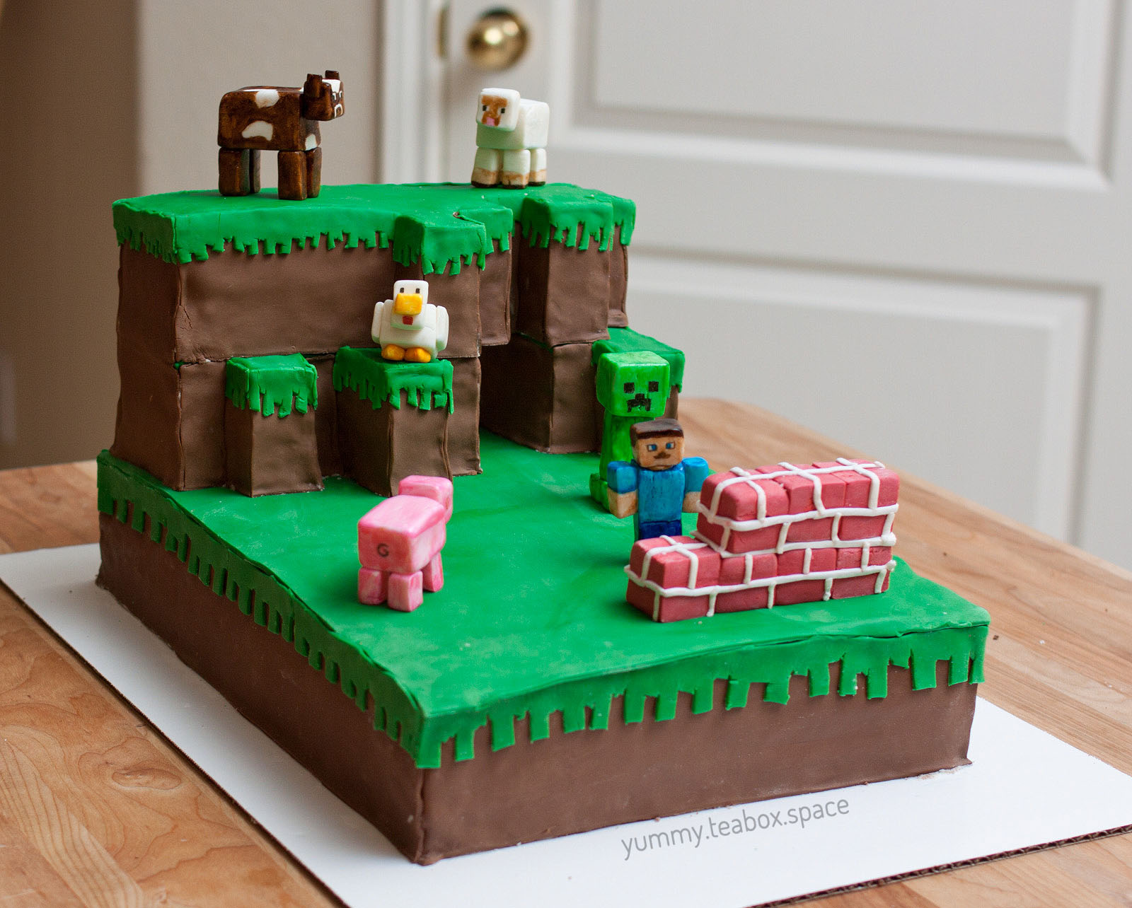 A cake that looks like a Minecraft scene with Steve and a Creeper behind him. There are Minecraft cow, sheep, chicken, and pig figures on the cake too.