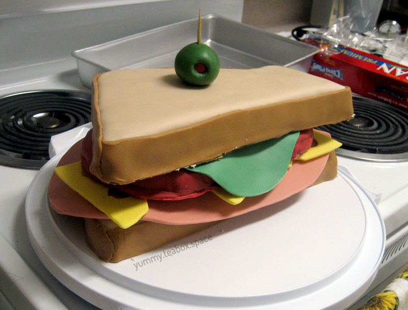 Cake that looks like the sandvich from Team Fortress 2, which is a triangular sandwich with meat, cheese, tomatoes, lettuce, and an olive on a toothpick.