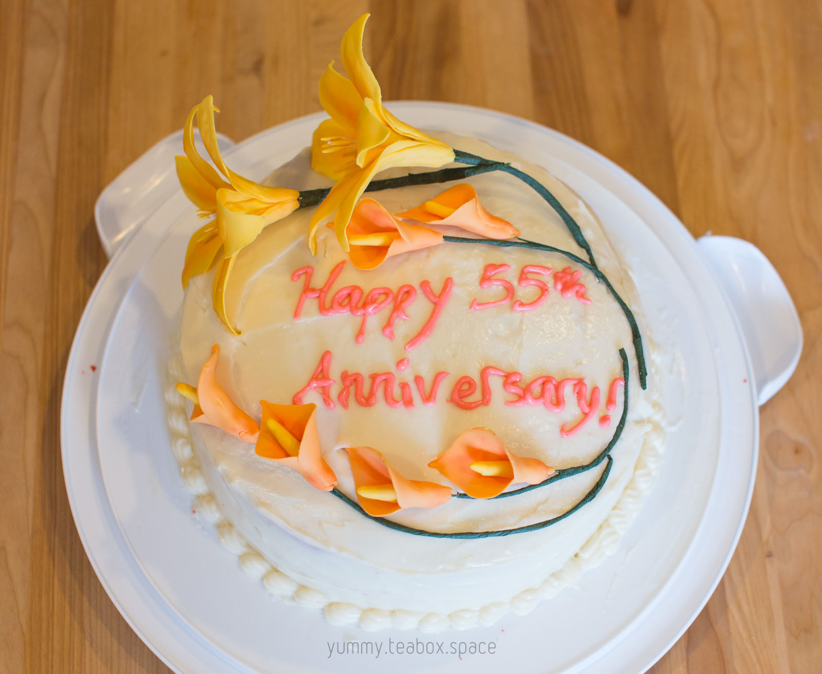 Round cake with white frosting and decorated with yellow lilies and orange calla lilies. The center says Happy 55th Anniversary.