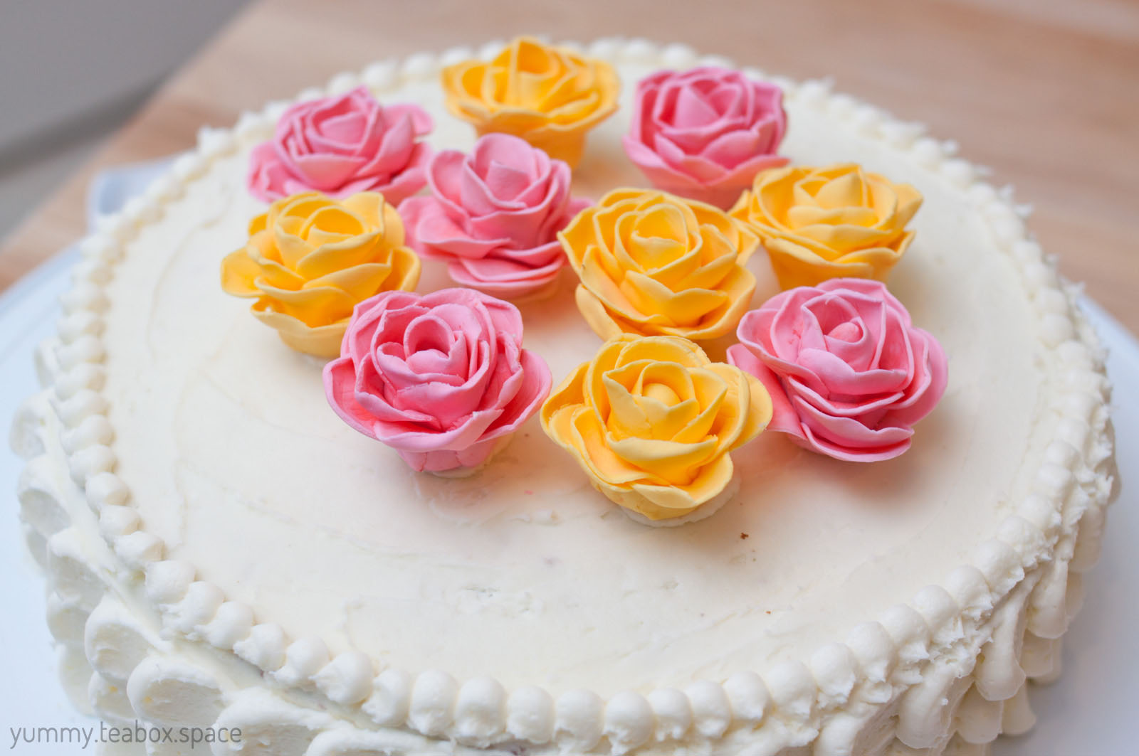Round cake with white frosting and yellow and pink roses in the center. The edges are in a scalloped pattern.