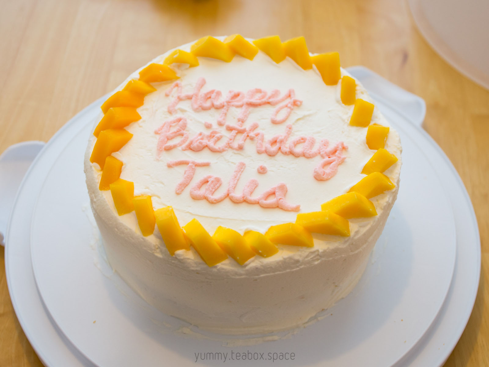 Round cake with whipped cream frosting and a border made with mango. The center says Happy Birthday Talia.