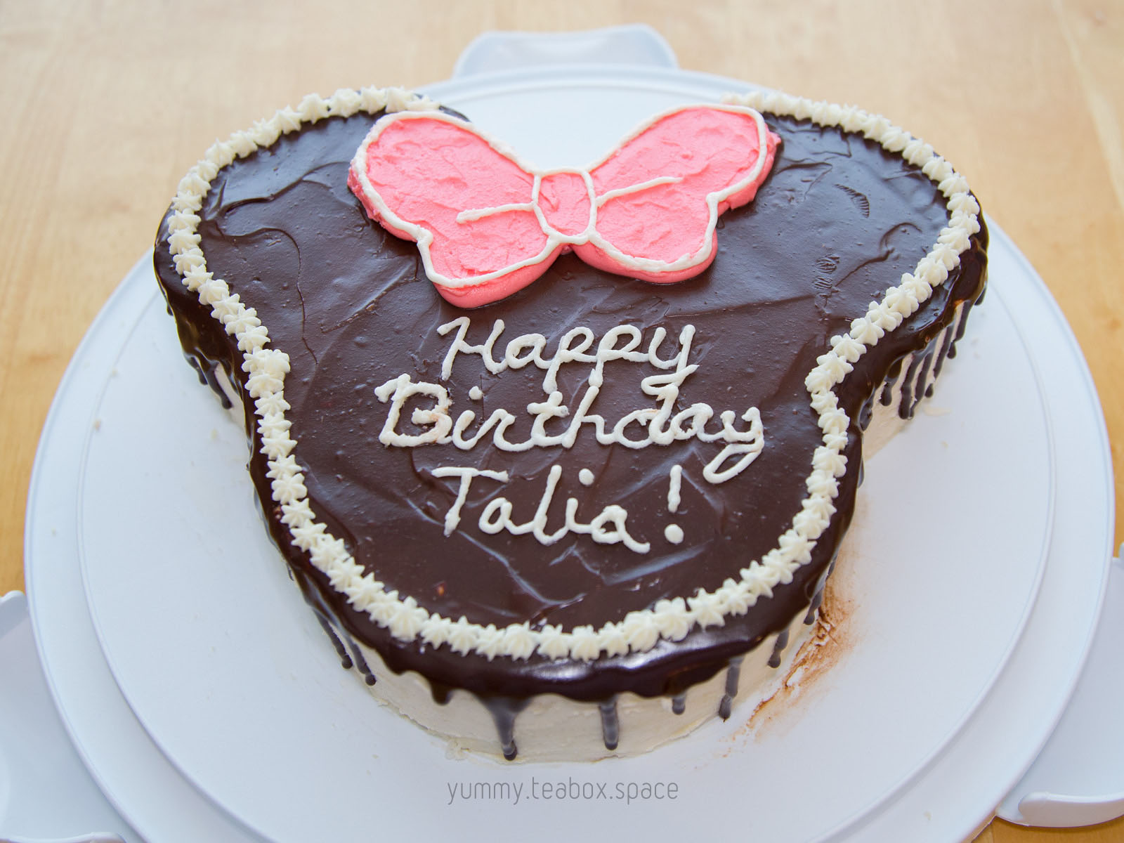 Cake shaped like Minnie Mouse's head, covered in a chocolate ganache drip. It has a pink bow, a white frosting border, and says Happy Birthday Talia.
