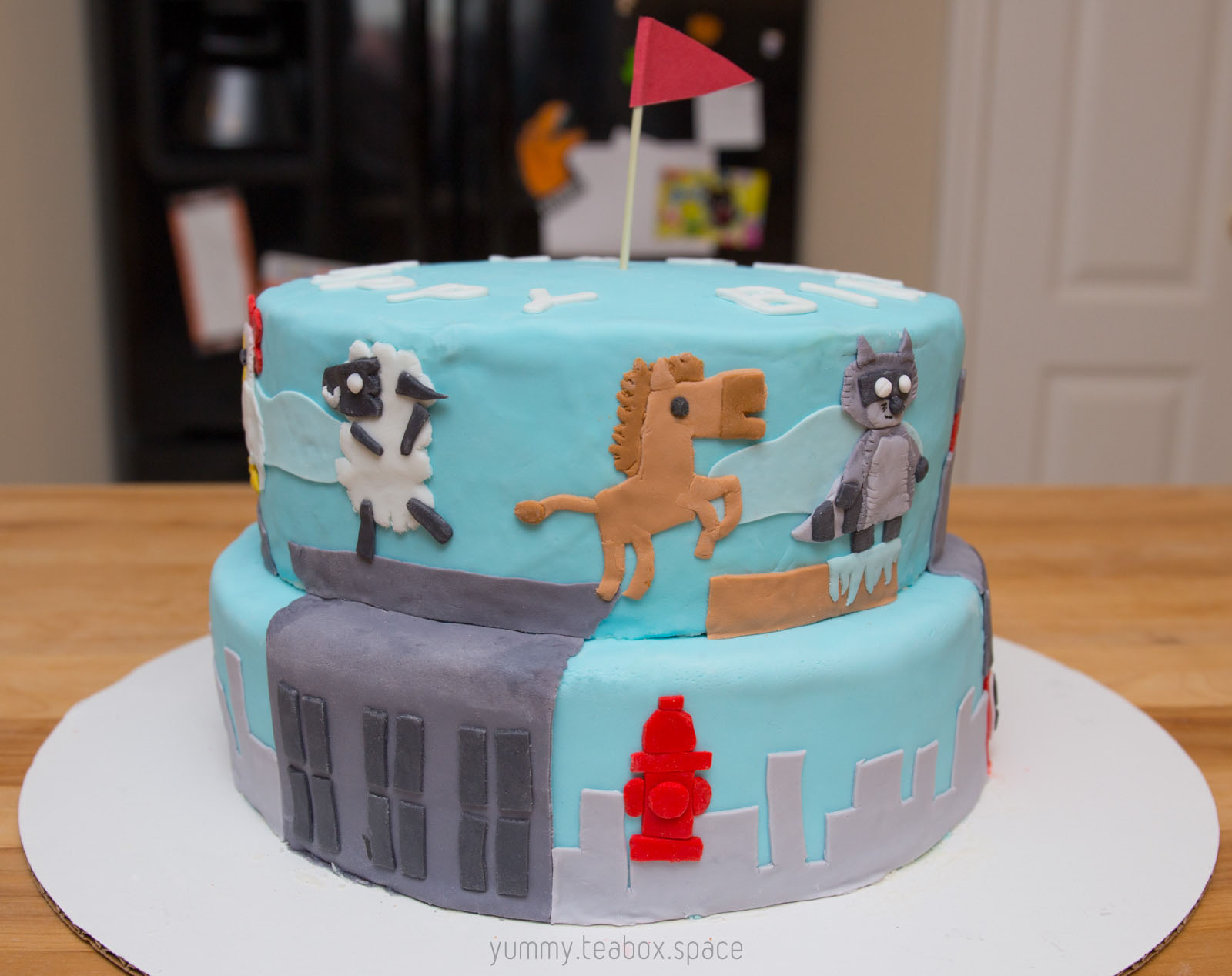2-tier blue cake with images of sheep, horse, and raccoon from Ultimate Chicken Horse on the side.