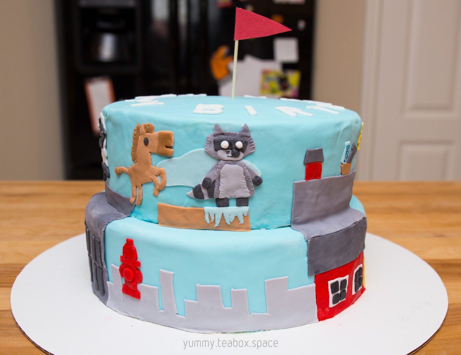 2-tier blue cake with images of horse and raccoon from Ultimate Chicken Horse on the side.
