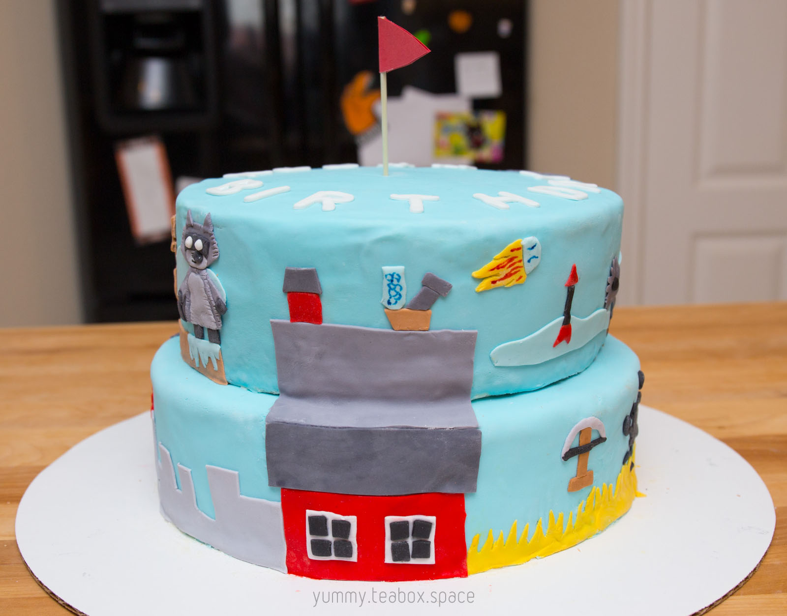 2-tier blue cake with images of racoon and traps on a red barn from Ultimate Chicken Horse on the side.