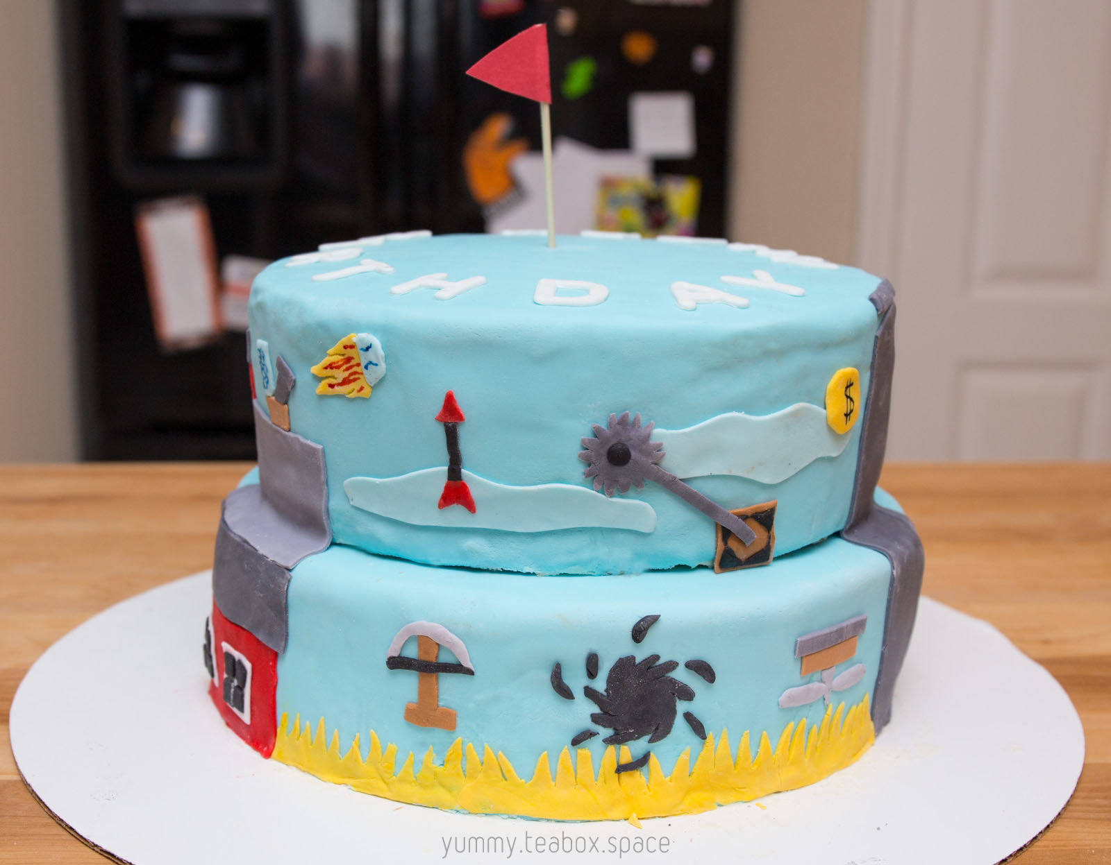 2-tier blue cake with images of traps from Ultimate Chicken Horse on the side.
