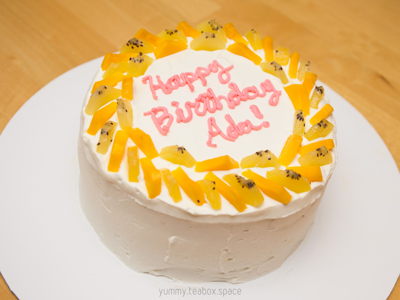 Round white cake with whipped cream frosting and a border of kiwi and mango. The center says Happy Birthday Ada.