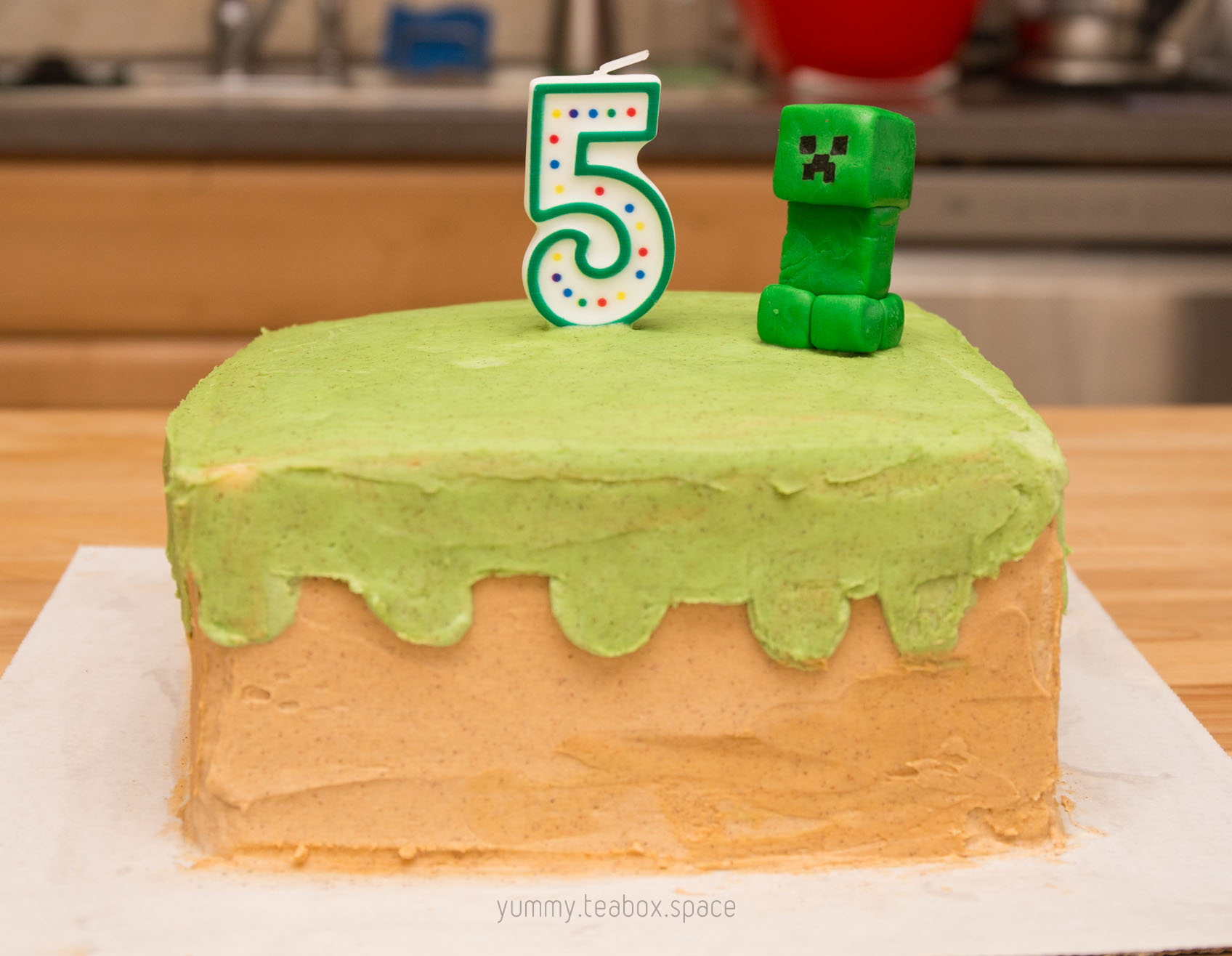 Cake that looks like a Minecraft grass block with brown dirt on the bottom and green grass on top. A Creeper sits on top with a 5 candle.
