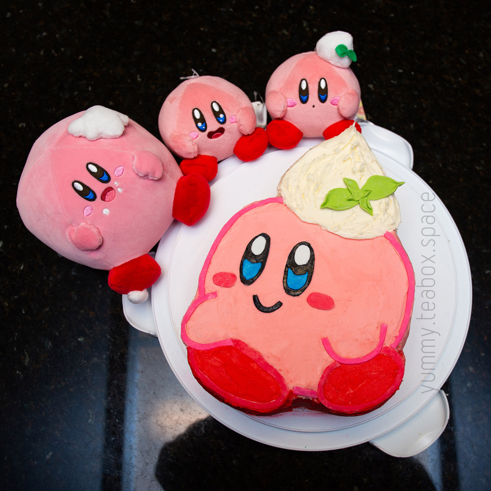 Top down view of a cake that looks like Kirby with a dollop of whipped cream on his head, with 3 Kirby plushies surrounding it.